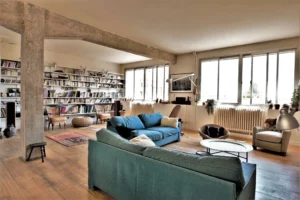 Espace de vie lumineux d’un loft parisien avec grande bibliothèque murale, canapés en tissu bleu, parquet ancien et fenêtres plein sud, à deux pas du marché Saint-Quentin.