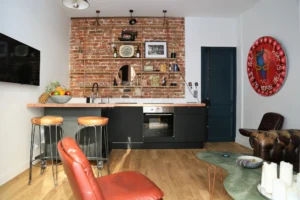 Cuisine d’un appartement à Montmartre Paris 18 avec mur en briques apparentes, étagères décoratives en cuivre, îlot central et fauteuil vintage en cuir.