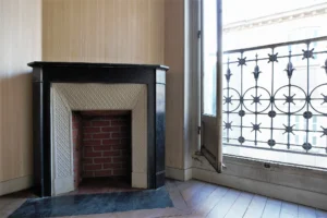 Détail d’une cheminée ancienne en faïence et bois noir avec intérieur en briques, à côté d’une fenêtre ouverte sur un garde-corps en fer forgé, dans un appartement ancien à Belleville Paris 19e.