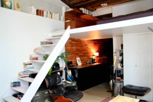 Vue sur l’escalier blanc menant à la mezzanine, bibliothèque, fauteuil vintage et mur en bois soulignant le caractère unique du lieu.