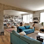 Vue intérieure d’un loft à Paris 10ᵉ avec poutres apparentes, grande bibliothèque, canapés turquoise et lumière naturelle orientée sud rue de Chabrol.
