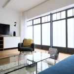 Coin salon cosy et lumineux, avec verrière atelier, fauteuil design, table basse en verre et sol en bois clair dans un loft à Paris 10.