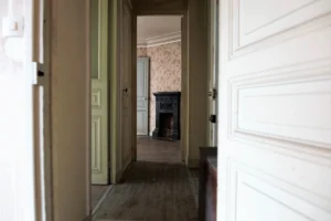 Vue d’un couloir ancien avec parquet et portes d’époque, menant à une pièce ornée d’une cheminée décorative en fonte, dans un appartement à rénover situé à Paris 19e, quartier Belleville.