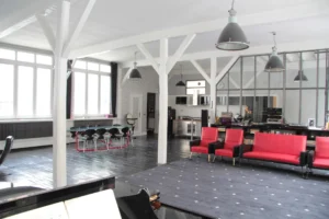 Vue d’ensemble sur un loft parisien avec cuisine ouverte, coin salle à manger, salon rétro, grande verrière atelier et sol en parquet noir, quartier Faubourg Saint-Denis.