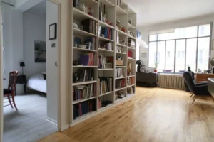 Vue sur la bibliothèque murale et l’entrée d’une chambre ouverte dans un loft lumineux à Paris 10ᵉ, avec parquet en bois clair et grande verrière.