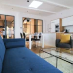 Vue en perspective du salon vers la cuisine, avec canapé bleu, verrière intérieure et mobilier contemporain dans un loft à Paris 10.