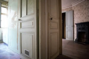 Vue croisée sur deux pièces séparées par des portes anciennes, avec à gauche un début de cuisine à rénover et à droite une chambre avec cheminée en fonte, dans un appartement ancien à Belleville, Paris 19e.