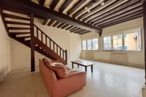 Salon lumineux d’un appartement à vendre à Pont-l’Évêque, avec escalier en bois, poutres apparentes et grandes fenêtres donnant sur la rue.