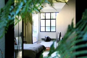 Chambre baignée de lumière naturelle, dissimulée derrière des feuillages, avec verrière atelier, rideaux fluides et ambiance douce.
