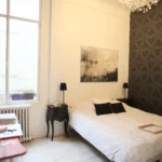 Chambre du loft avec sol en parquet clair, grande fenêtre, lustre en cristal et mur décoré de papier peint graphique à Paris 10ᵉ.