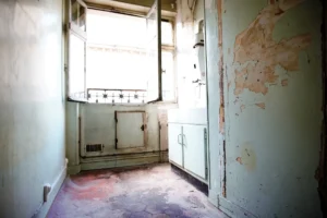 Cuisine ancienne à rénover avec murs à reprendre, sol en tomettes usées et vue dégagée sur cour intérieure, dans un appartement situé à Paris 19e, quartier Belleville.