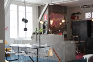 Salle à manger d’un loft parisien avec table noire, chaises minimalistes, cuisine ouverte en béton et suspensions design dans une ambiance lumineuse et éclectique.
