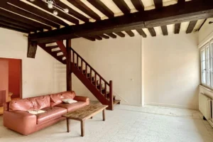 Salon d’un appartement à vendre à Pont-l’Évêque, avec plafond à poutres apparentes, escalier en bois massif et canapé en cuir.
