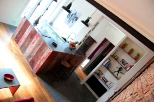 Vue d’un îlot central en bois brut avec corbeille de fruits, suspensions noires, miroir doré et mur en briques dans un appartement à Belleville – Paris 19ᵉ.