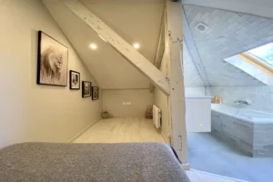 Espace nuit sous combles avec poutres apparentes et accès direct à la salle de bains dans un duplex à Deauville.