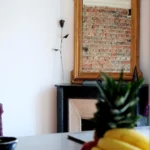 Reflet du mur en briques dans un miroir doré au-dessus d’une cheminée noire, dans un appartement à vendre à Paris 19ᵉ.