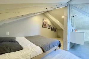 Suite mansardée avec lit double, poutres apparentes et salle de bains attenante dans un appartement duplex à Deauville.