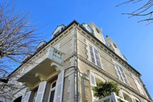 Façade d’un immeuble ancien typique de Deauville avec volets blancs et balcon, abritant un appartement duplex.