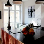 Cuisine lumineuse avec îlot central en bois patiné, grande fenêtre et suspensions noires, dans un appartement à Belleville – Paris 19ᵉ.