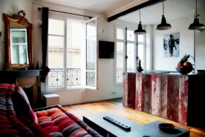 Espace salon-cuisine lumineux avec grande baie vitrée, canapé coloré et îlot central en bois patiné, dans un appartement à vendre à Paris 19ᵉ – quartier Belleville.