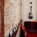 Mur en briques avec tableaux en noir et blanc, banquette colorée et guitare suspendue dans un salon d’appartement à Belleville – Paris 19ᵉ.
