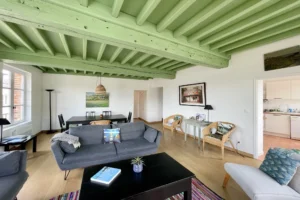 Salon convivial avec canapé gris, lumière traversante et accès direct à la cuisine dans un appartement en centre-ville de Honfleur.