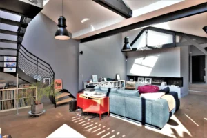 Vue du salon dans un loft à Nanterre avec escalier en métal, poutres apparentes et décoration de style industriel.