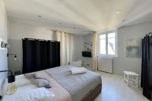 Chambre lumineuse avec lit double, télévision murale et fenêtre donnant sur les toits de Deauville dans un appartement duplex.