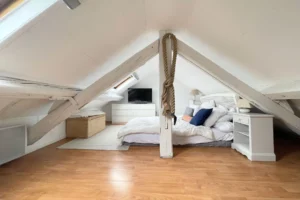 Chambre en mezzanine sous les toits avec lit double, poutres blanches et parquet.