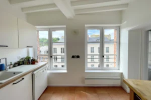 Cuisine baignée de lumière avec vue dégagée sur les toits du centre historique d’Honfleur, dans un appartement à vendre.