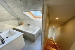 Salle de bains mansardée avec baignoire et vasque moderne, éclairée par une fenêtre de toit dans un duplex à Deauville.