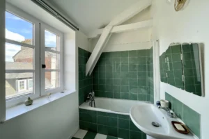 Salle de bains avec baignoire, carrelage vert et poutre apparente sous combles dans un appartement à vendre à Honfleur.