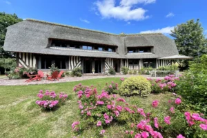 Maison normande à vendre à Ouilly-le-Vicomte avec toit de chaume, jardin fleuri et mobilier de jardin sous un ciel bleu, proche de Lisieux.