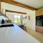 Cuisine équipée et ouverte sur l’extérieur, avec poutres en bois, mobilier contemporain et lumière naturelle à Ouilly-le-Vicomte.
