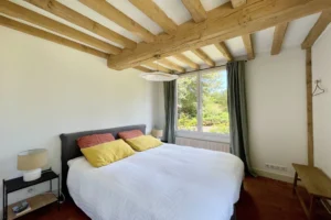 Chambre parentale avec poutres apparentes, lit double, grandes fenêtres et vue sur le jardin à Ouilly-le-Vicomte.