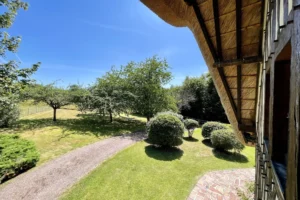 Vue depuis l’étage sur le jardin arboré, chemin en gravier et toit en chaume d’une maison à Ouilly-le-Vicomte.