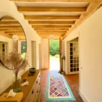 Entrée lumineuse d’une maison normande à Ouilly-le-Vicomte, avec poutres apparentes, carrelage ancien et tapis coloré.