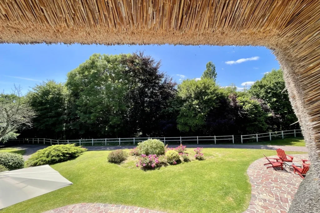 Vue depuis l’étage sous le toit en chaume, avec terrasse, jardin paysager et clôture en bois à Ouilly-le-Vicomte.