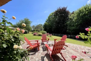 Terrasse en pierre avec fauteuils rouges, rosiers fleuris et vaste jardin arboré à Ouilly-le-Vicomte, sous un ciel bleu.