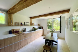 Cuisine lumineuse avec mobilier en bois clair, poutres apparentes et vue verdoyante sur le jardin à Ouilly-le-Vicomte.