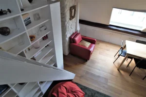 Vue depuis la mezzanine sur le salon avec fauteuil rouge, bibliothèque intégrée et espace repas sous combles, appartement rue de Turenne Paris 3ᵉ.