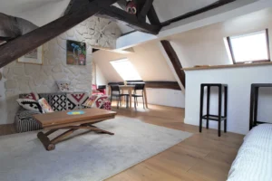 Coin salon et espace repas sous combles avec poutres apparentes et mur en pierres, table et chaises près d’une fenêtre, appartement rue de Turenne Paris 3ᵉ.