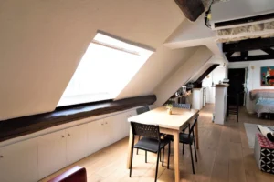 Espace repas sous combles avec table et chaises face à la cuisine ouverte, poutres apparentes et rangements intégrés, appartement rue de Turenne Paris 3ᵉ.