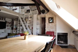 Espace bibliothèque avec mezzanine et cheminée sous combles, murs en pierres et poutres apparentes, appartement rue de Turenne Paris 3ᵉ.