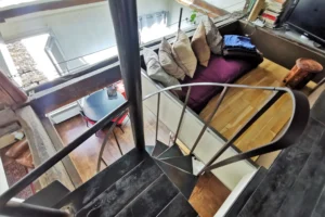 Mezzanine d’un loft à vendre à Paris 18, quartier Marx Dormoy – Marché de l’Olive, accessible par un escalier en métal et aménagée avec un coin détente sous poutres apparentes.