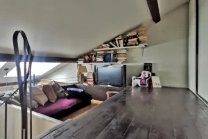 Espace détente aménagé en mezzanine dans un loft à vendre à Paris 18, quartier Marx Dormoy – Marché de l’Olive, avec bibliothèque, poutres apparentes et coin lecture cosy.