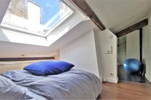 Chambre sous combles d’un loft à vendre à Paris 18, quartier Marx Dormoy – Marché de l’Olive, avec poutres apparentes, fenêtre de toit et lumière naturelle.