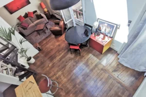 Vue en hauteur du séjour d’un loft à vendre à Paris 18, quartier Marx Dormoy – Marché de l’Olive, avec parquet ancien, coin salon et espace repas sous verrière.