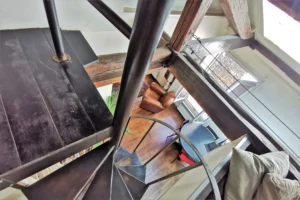 Escalier en colimaçon d’un loft à vendre à Paris 18, quartier Marx Dormoy – Marché de l’Olive, reliant la mezzanine au séjour avec vue sur le salon et les poutres apparentes.
