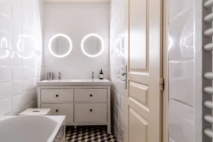 Salle de bains avec baignoire, double vasque et miroirs lumineux circulaires, carrelage blanc et sol géométrique, Paris 10 – Gare de l’Est.